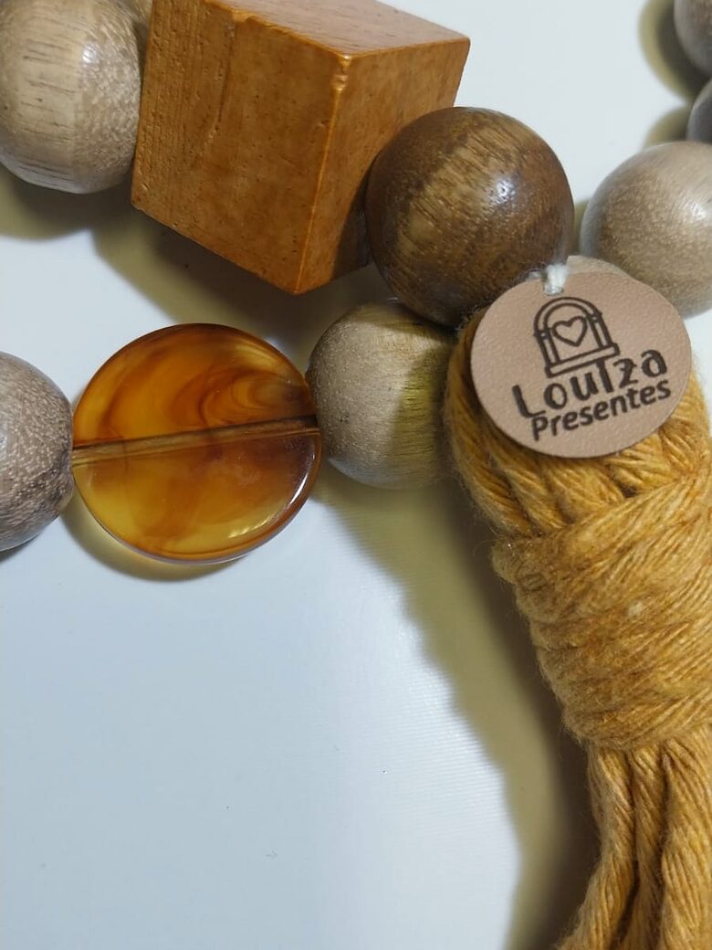 Detalhe de um colar de mesa: cubo de madeira, disco âmbar, borla mostarda e a etiqueta Louiza Presentes.