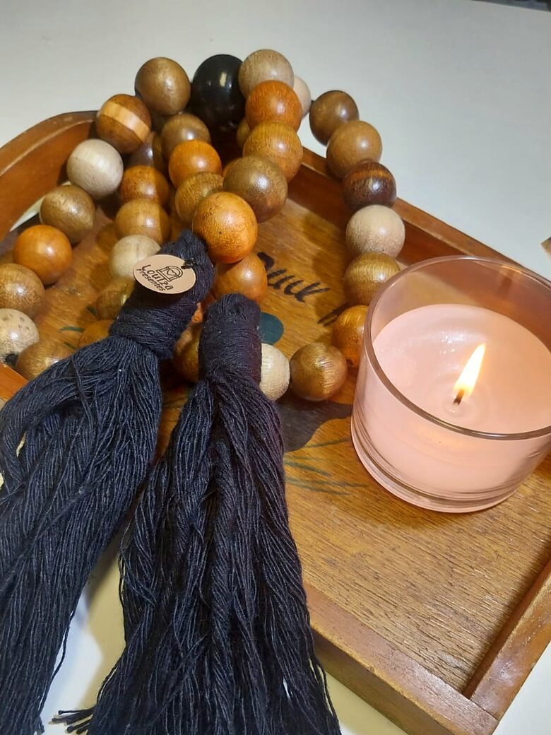 Colar de mesa em contas de madeira em tons de mel, com duas borlas pretas, sobre uma bandeja de madeira ao lado de uma vela acesa.